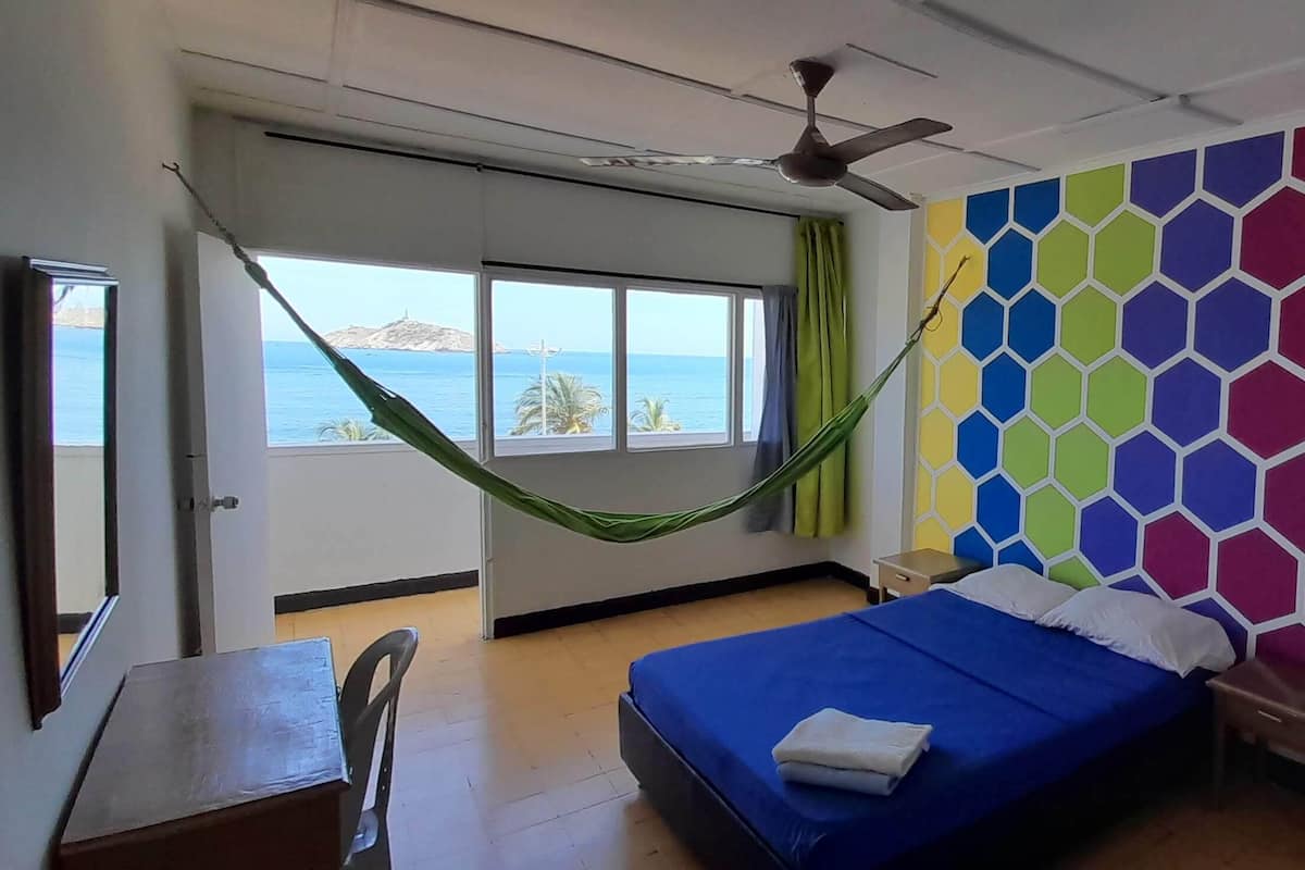 A bedroom with a bed, a desk, a chair, a hammock, a ceiling fan, and a view of the sea.