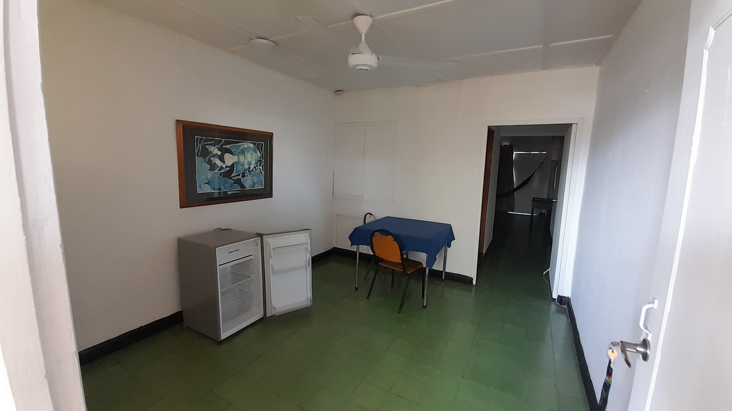 Une petite pièce propre, équipée d’un bureau, d’une chaise et d’un ventilateur de plafond.