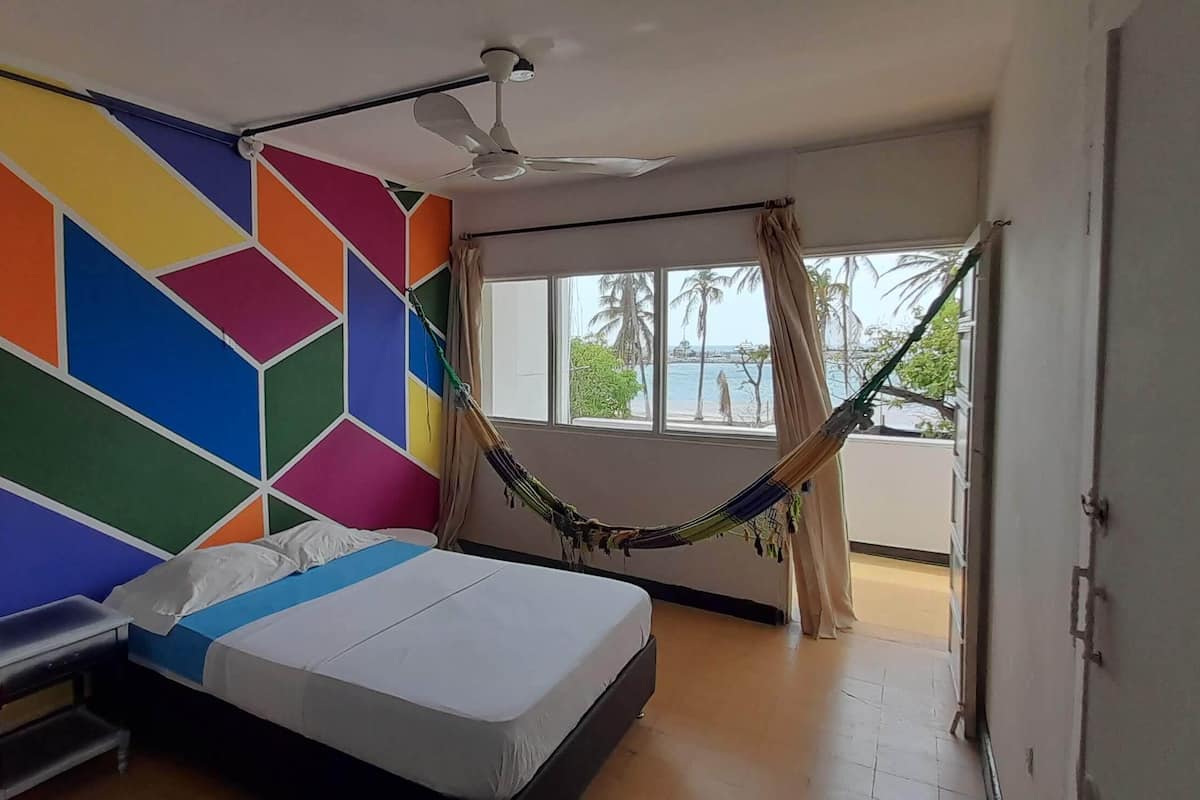 A bedroom with a colorful geometric wall design, a bed with a blue and white bedspread, a ceiling fan, and a hammock near a window with a view of palm trees and the sea.