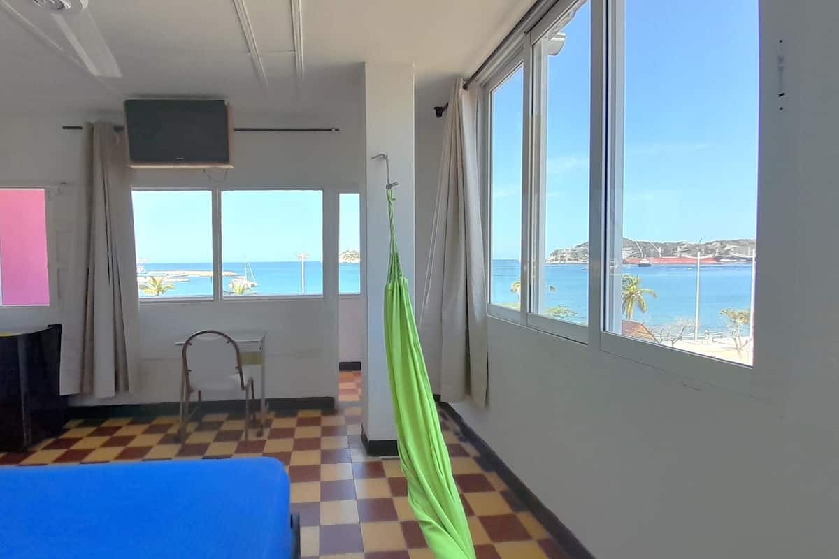 A room with a checkered floor, a bed, a TV, and a view of the sea through large windows.