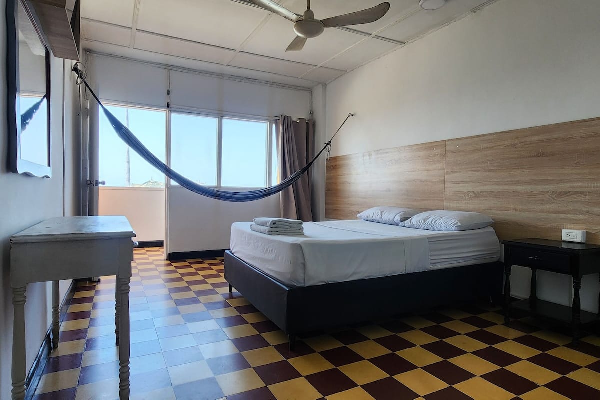 A hotel room with a checkered floor, a bed with a wooden headboard, a nightstand, a desk, a mirror, and a hammock.