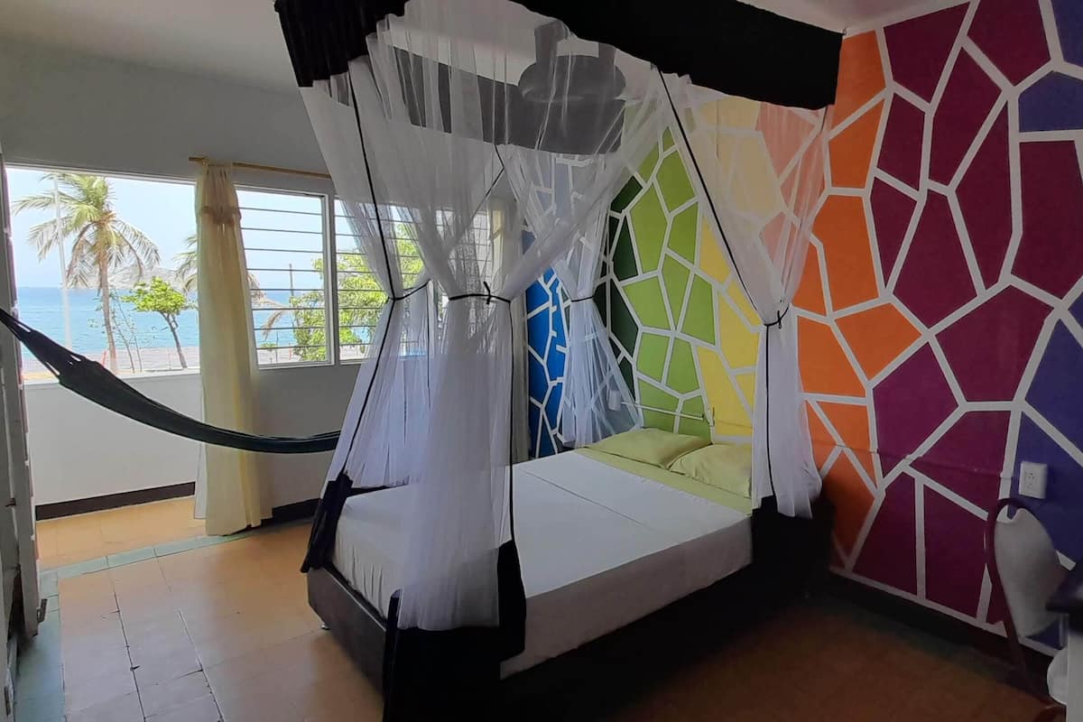 A room with a bed, a colorful geometric wall, a view of palm trees and the sea through a window, and a mosquito net over the bed.
