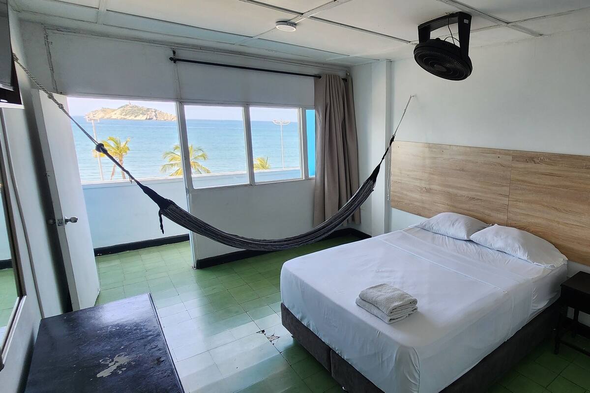 A hotel room with a bed, a nightstand, a hammock, a window with a view of the sea, and a ceiling fan.