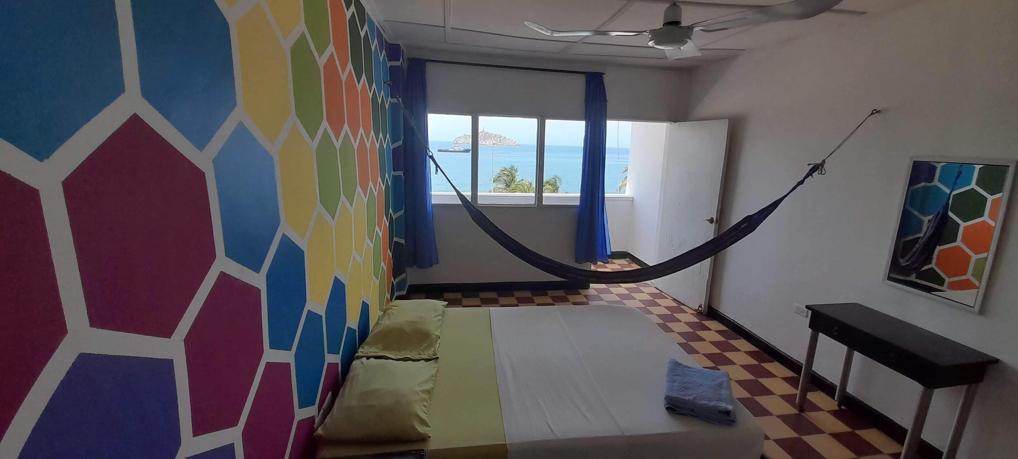 Een kamer met een hangmat, een bed, een zwart-wit geruite vloer, een raam met uitzicht en een kleurrijke muur.