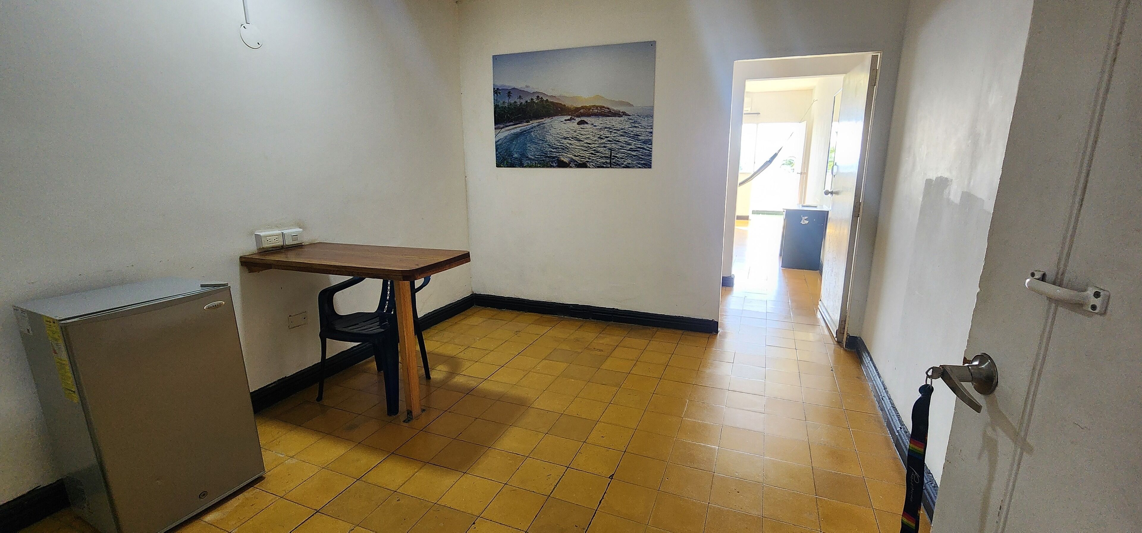A small room with a wooden table, a black chair, a small refrigerator, and a painting of a coastal scene on the wall.