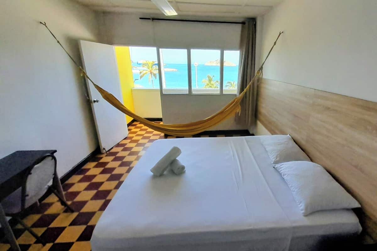 A hotel room with a bed, a checkered floor, a hammock, and a view of the ocean through a window.