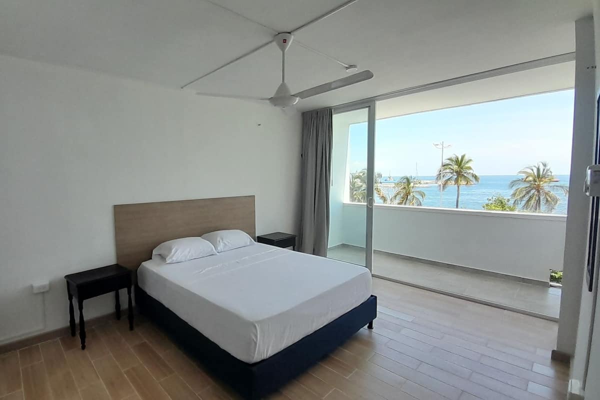 A bedroom with a bed, a nightstand, a ceiling fan, and a view of the ocean through a balcony.
