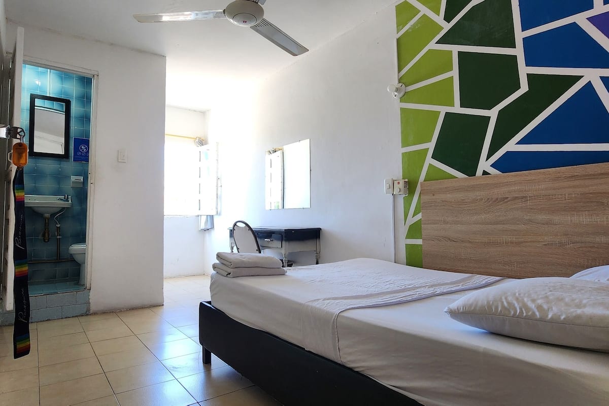 A bedroom with a bed, a ceiling fan, a bathroom with a toilet and sink, and a colorful wall design.
