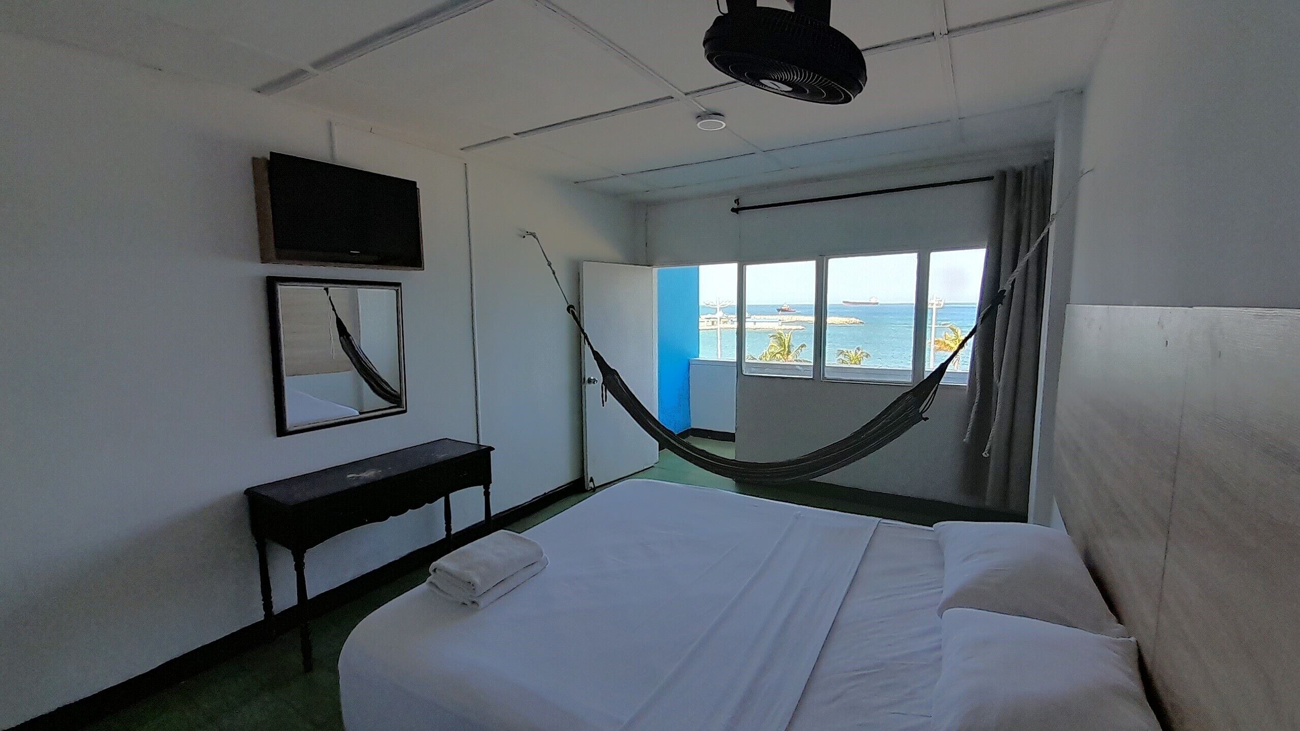 A hotel room with a bed, a desk, a TV, a hammock, and a view of the sea.