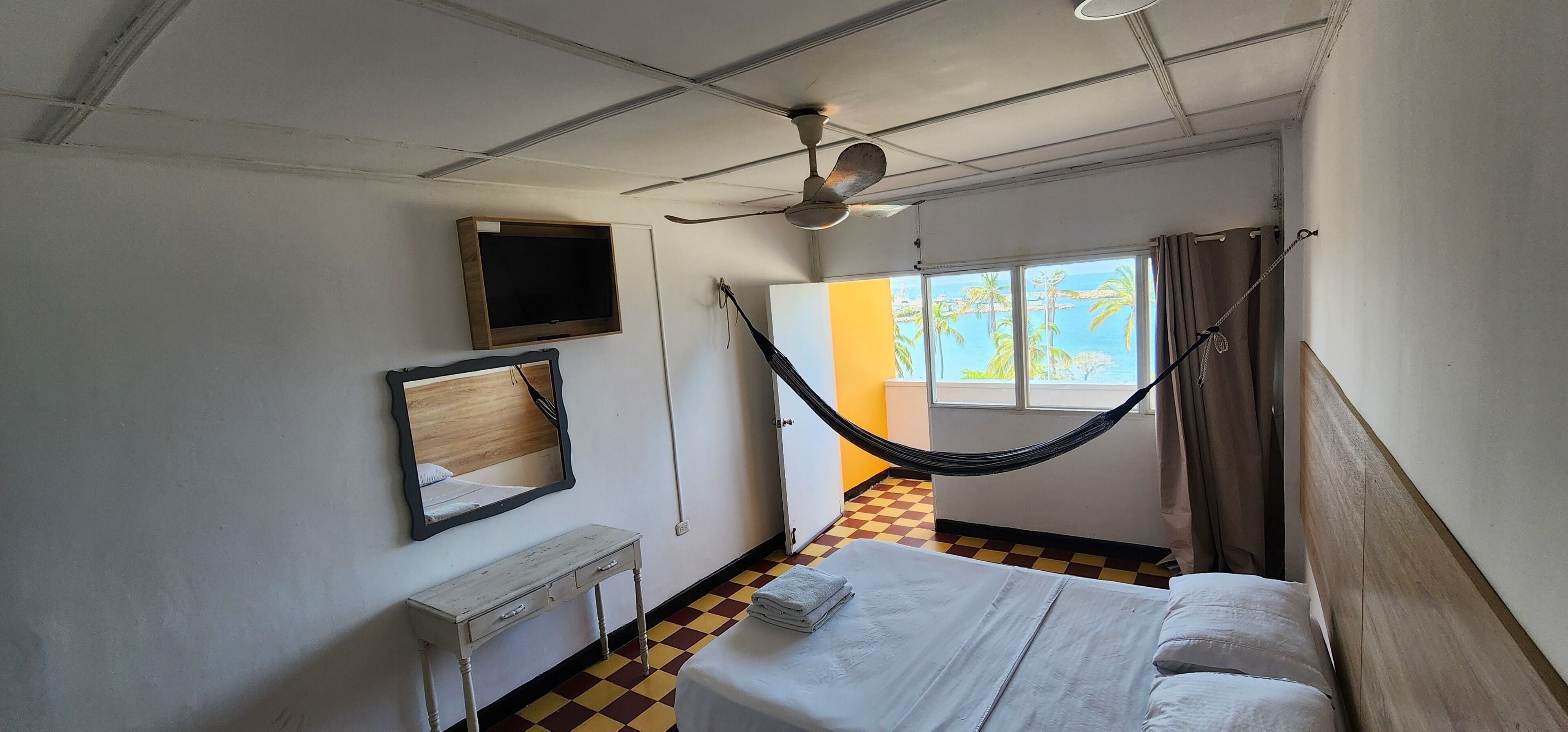 Une chambre d’hôtel équipée d’un lit, d’un bureau, d’un miroir, d’un ventilateur de plafond et d’une fenêtre donnant sur des palmiers.