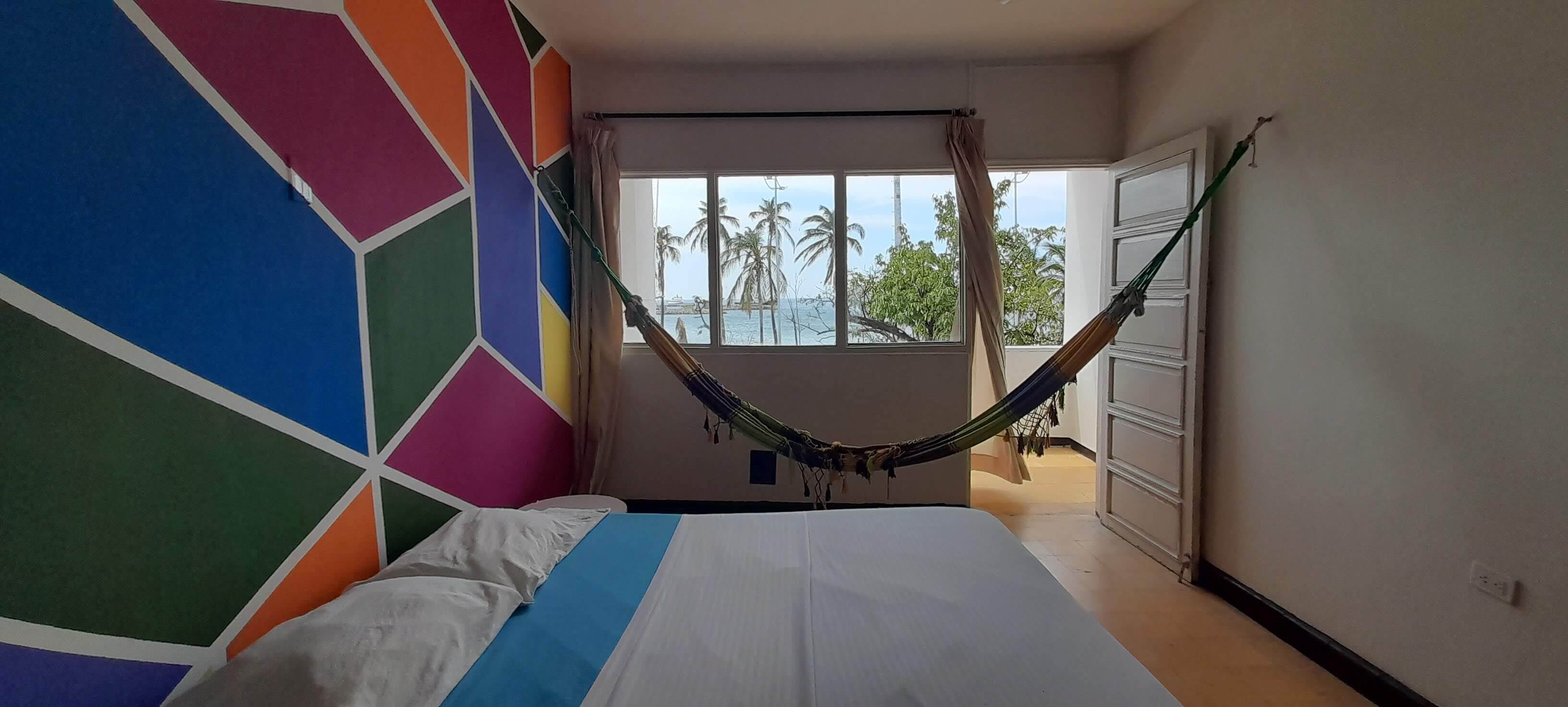 Een slaapkamer met een hangmat, een wand met kleurrijke geometrische patronen, een bed met wit- en blauw beddengoed, en uitzicht op palmbomen door een raam.