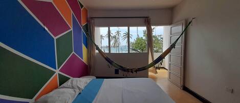 Een slaapkamer met een hangmat, een wand met kleurrijke geometrische patronen, een bed met wit- en blauw beddengoed, en uitzicht op palmbomen door een raam.