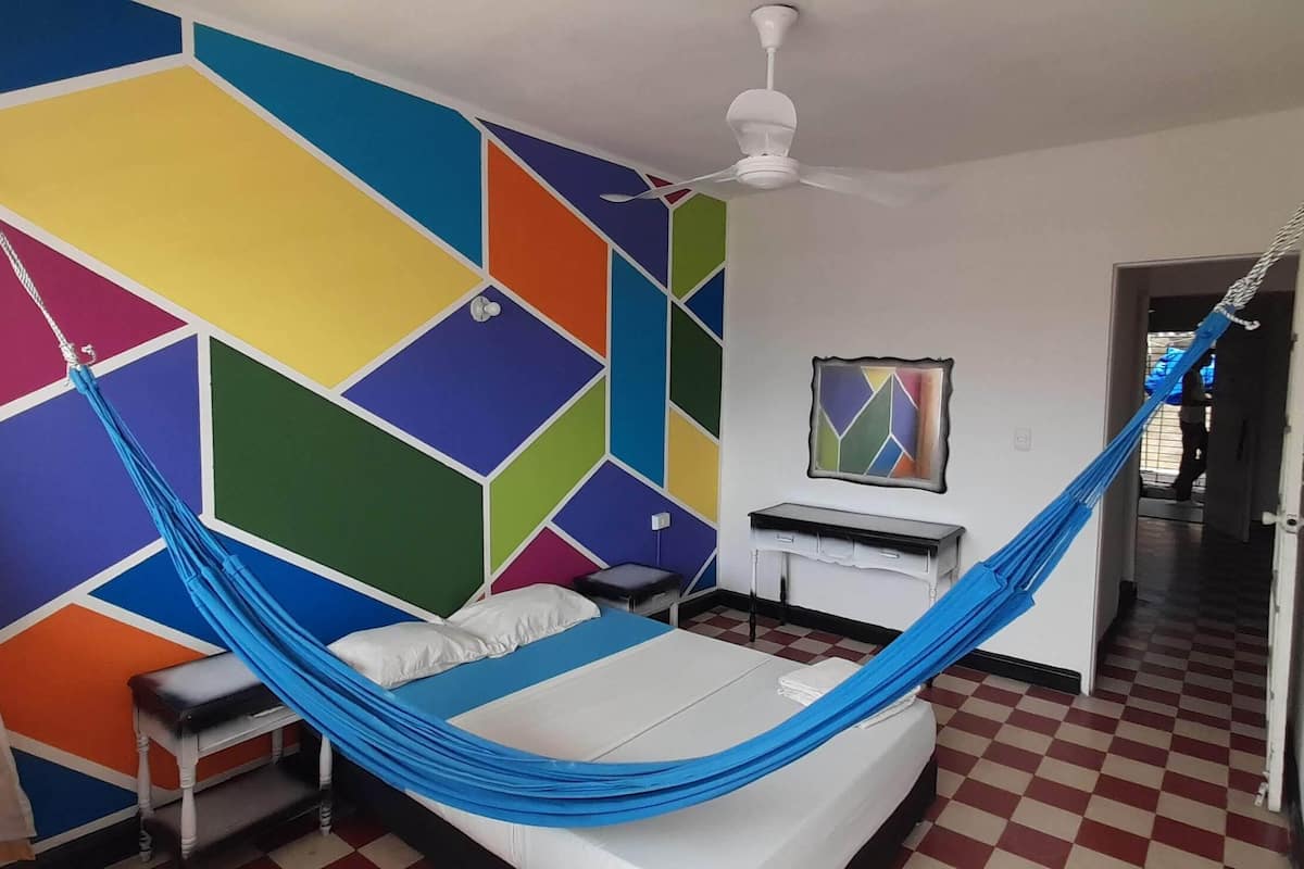 A room with a bed, a hammock, a ceiling fan, a desk, and a colorful geometric wall.