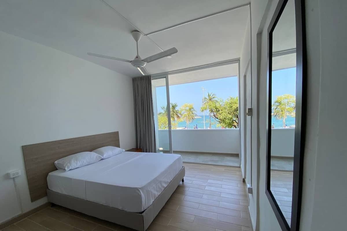 A hotel room with a bed, a ceiling fan, and a balcony with a view of palm trees and a pool.