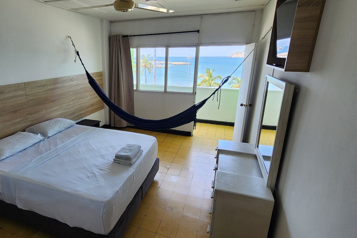 A hotel room with a bed, a bedside table, a small cabinet, a hammock, and a view of the sea through a window.