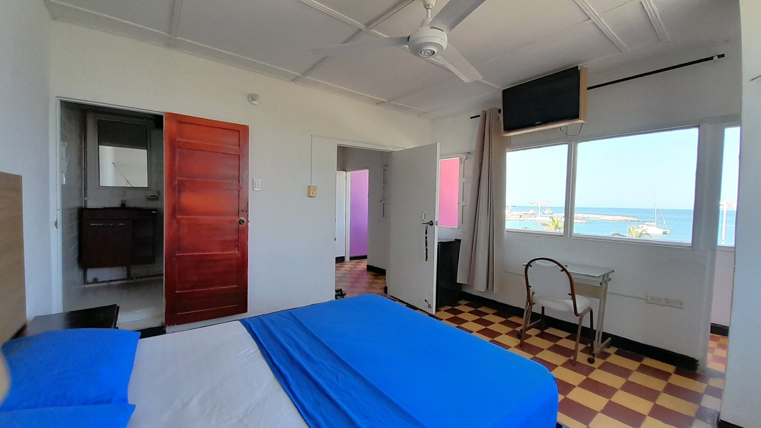 A hotel room with a bed, a TV, a window with a view of the sea, and a ceiling fan.