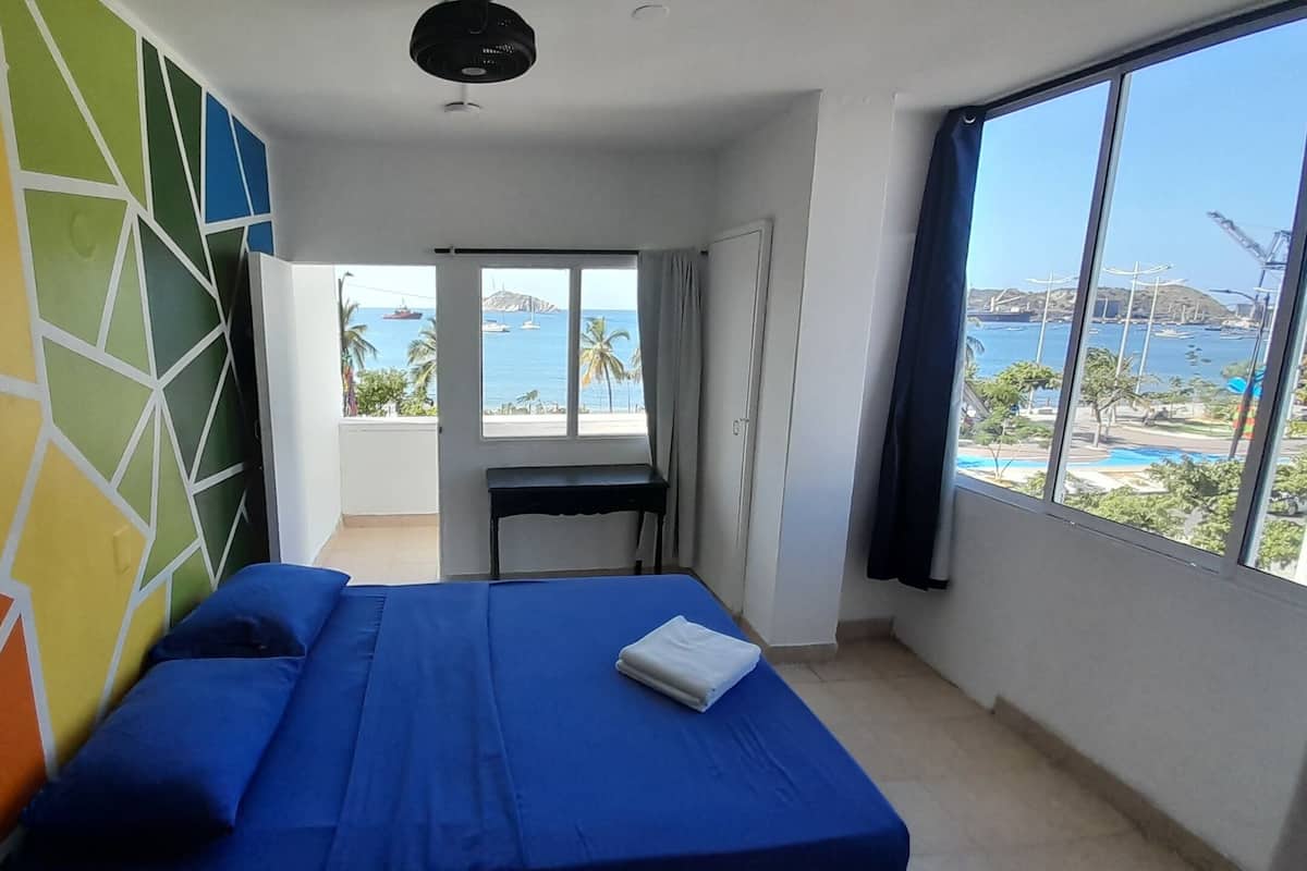 A room with a bed, a desk, and a large window with a view of the sea and boats.