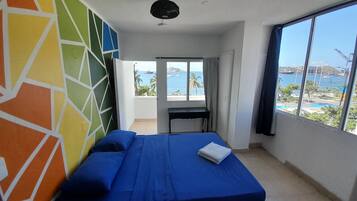 Una habitación con cama, escritorio y un amplio ventanal con vistas al mar y a los barcos.