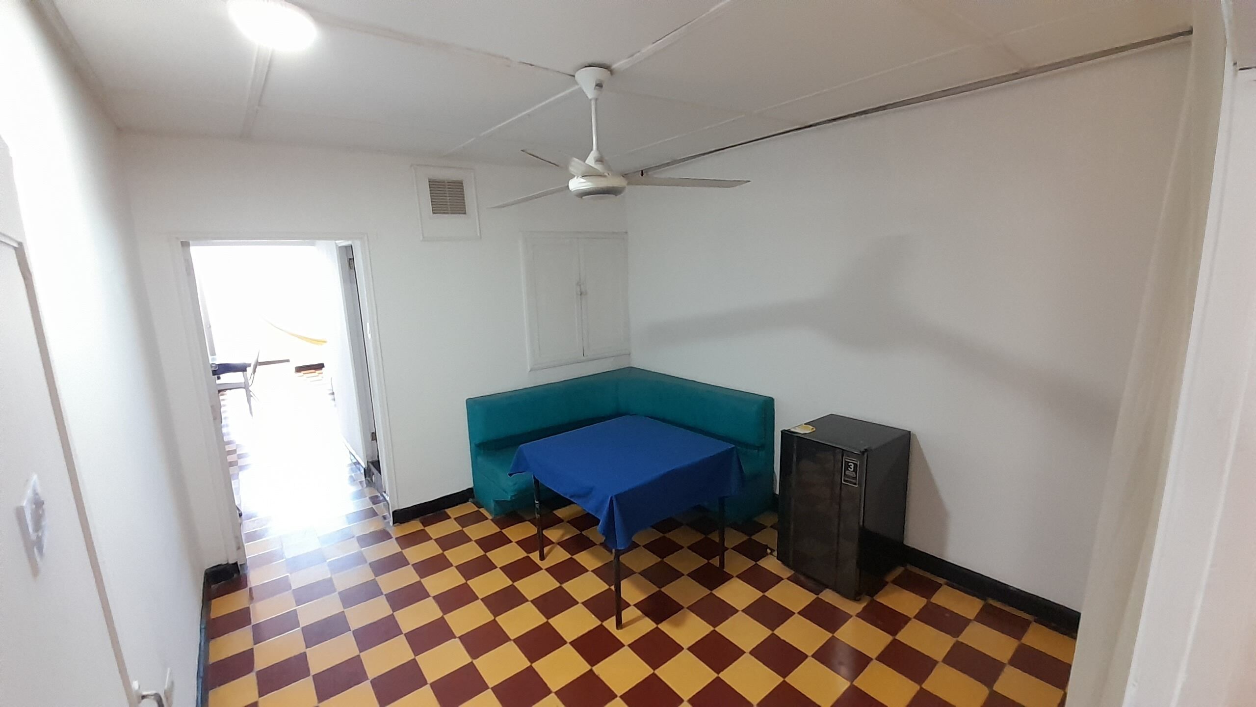 Une pièce avec un sol à carreaux, un ventilateur de plafond, une table bleue, un canapé turquoise et un petit meuble noir.