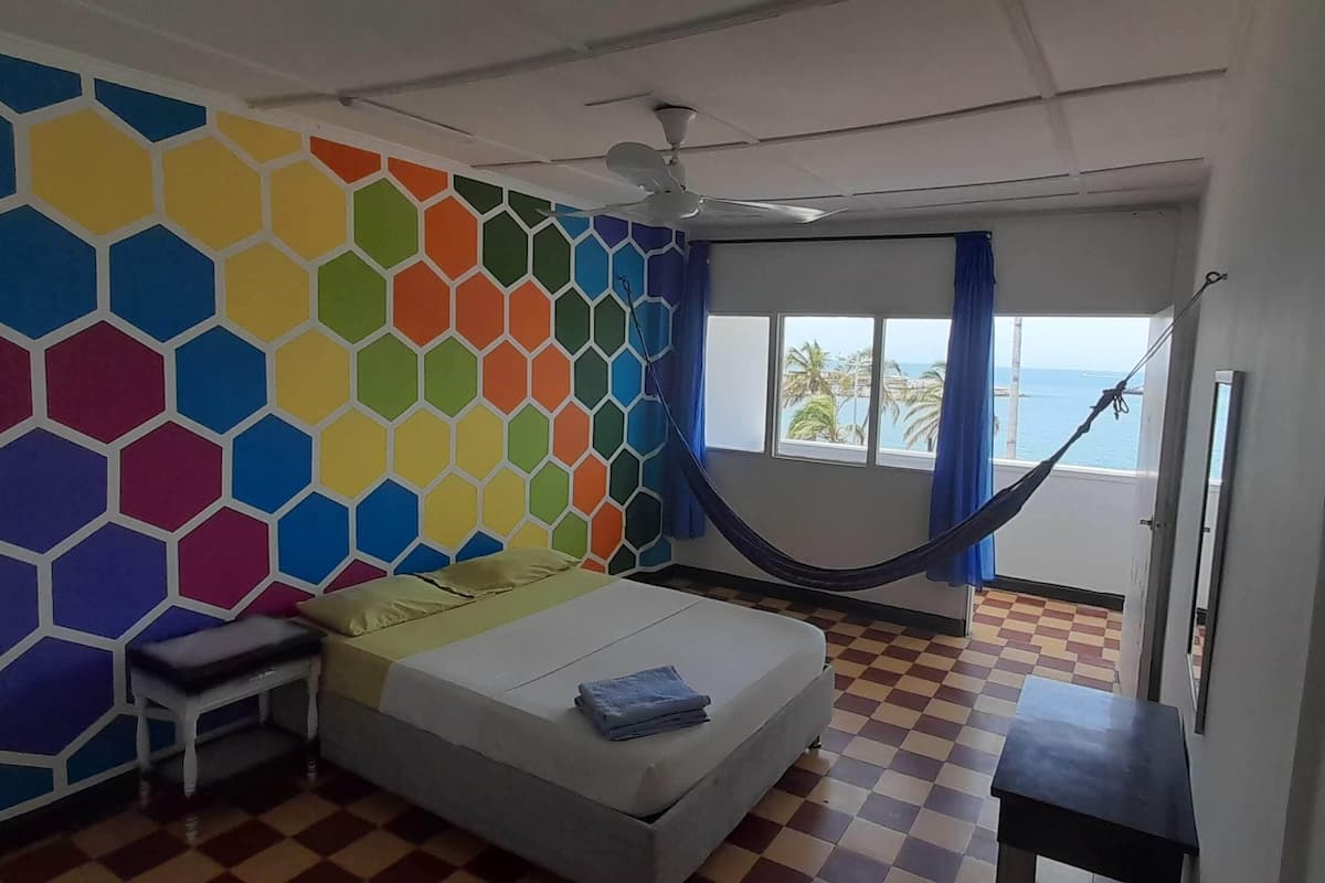 A room with a bed, a small table, a chair, a hammock, a window with a view of palm trees, and a colorful hexagonal wall design.
