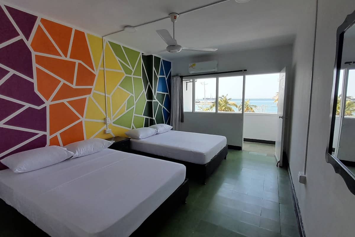 A hotel room with two beds, a colorful geometric wall, a ceiling fan, and a large window with a view of the sea.