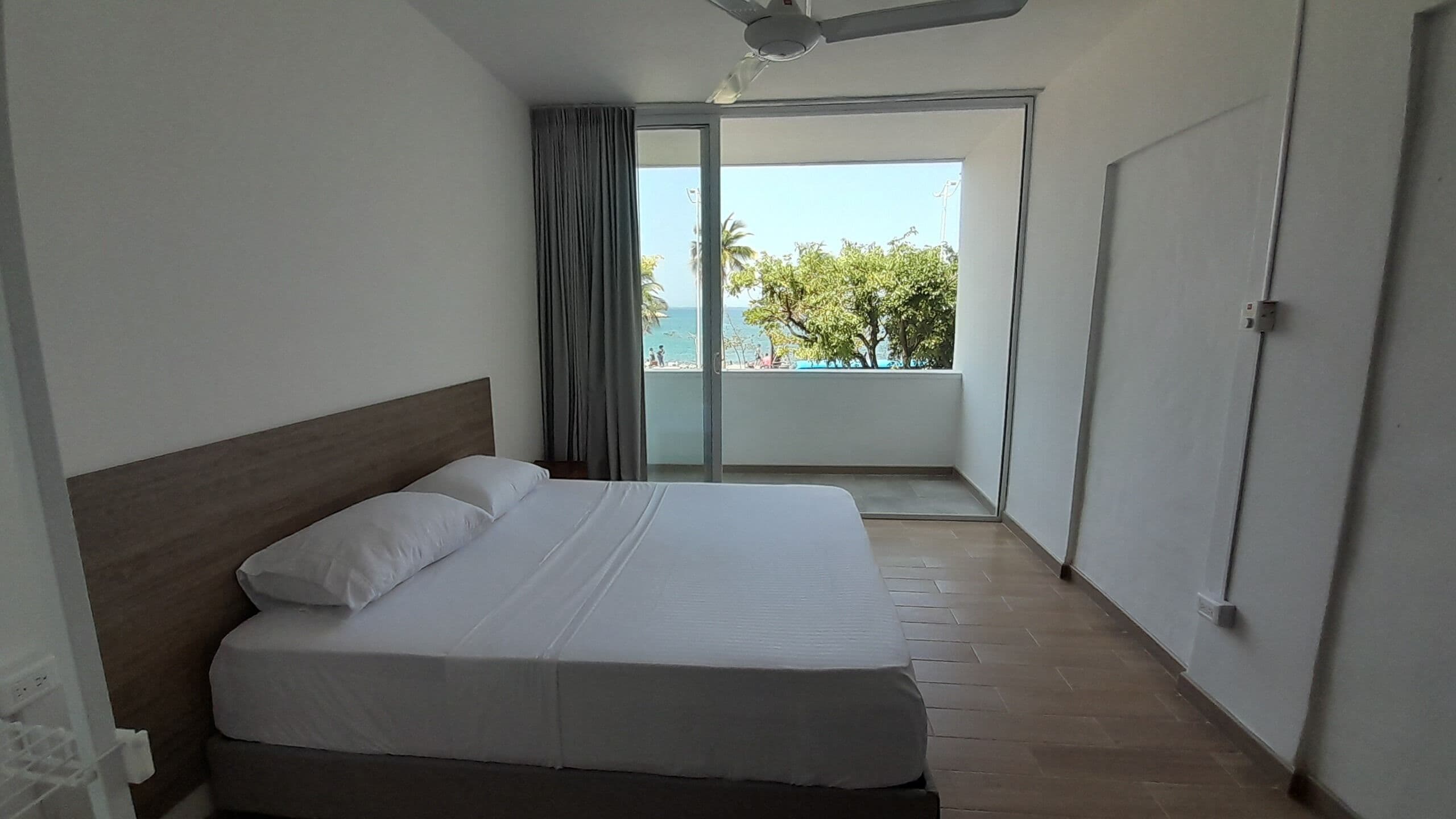 Habitación de hotel con cama, ventilador de techo, vista al mar y balcón con puerta.