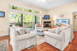 A living room with two white sofas, a coffee table, a fireplace, a television, and a large sliding glass door leading to a balcony.
