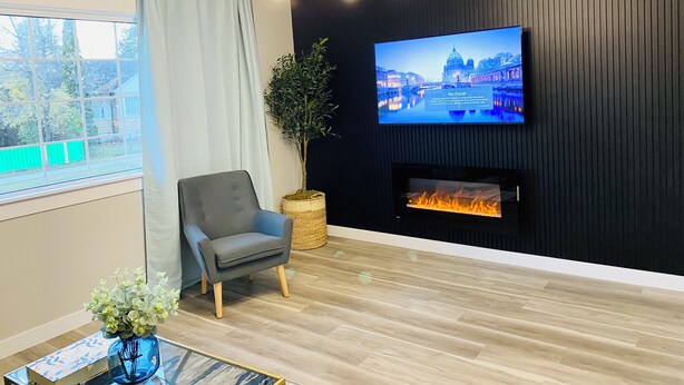 Ett modernt vardagsrum med en tv monterad på en mörk vägg, en öppen spis, en bekväm fåtölj och utsikt över grönområden utanför.