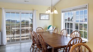 A dining room with a large wooden table, six chairs, a chandelier, and large windows with a view of the outdoors.