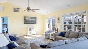 A living room with a ceiling fan, a large window with blinds, a television, and a view of the outdoors.