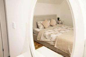 Een slaapkamer met een bed, kussens, een nachtkastje met een plant en een klein nachtkastje.