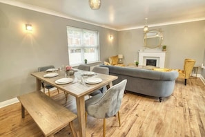 Een eetgedeelte met een houten tafel voor vier personen, een grijze bank en een open haard.