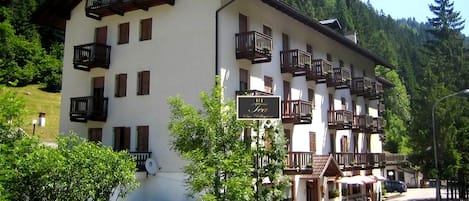 Un hotel de varios pisos con balcones, un cartel que indica 'HT Tea San Pellegrino' y un paisaje montañoso de fondo.