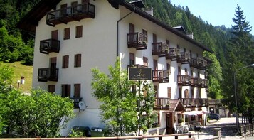 Un hotel de varios pisos con balcones, un cartel que indica 'HT Tea San Pellegrino' y un paisaje montañoso de fondo.