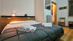 A neatly made bed with a dark green quilt and rolled towels, a wooden headboard, a bedside lamp, a radiator, a chair, and a mirror on the wall. - Ht TEA San Pellegrino (Canale d'Agordo)