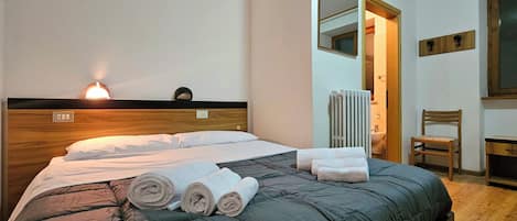 Una cama bien hecha con colcha verde oscuro y toallas enrolladas, cabecero de madera, lámpara de noche, radiador, una silla y un espejo en la pared.