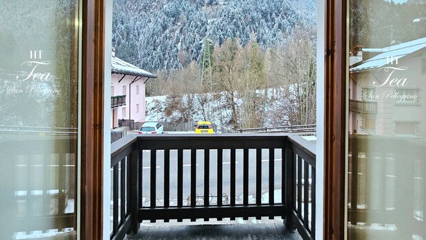 A wooden balcony with a view of a snow-covered mountain and a yellow car parked on the street below. - Ht TEA San Pellegrino (Canale d'Agordo)