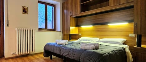 Un dormitorio con cama, cabecera de madera, ventana, radiador y un pequeño cuadro en la pared.