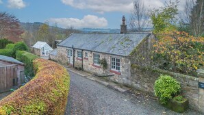A stone building with a sloped roof, a chimney, and a small garden area with a hedge and a bush.
