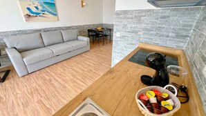 Ein modernes Wohnzimmer mit grauem Sofa, einem Essbereich mit Tisch und Stühlen sowie einer Küche mit Spüle und einem Korb mit Wasserflaschen.