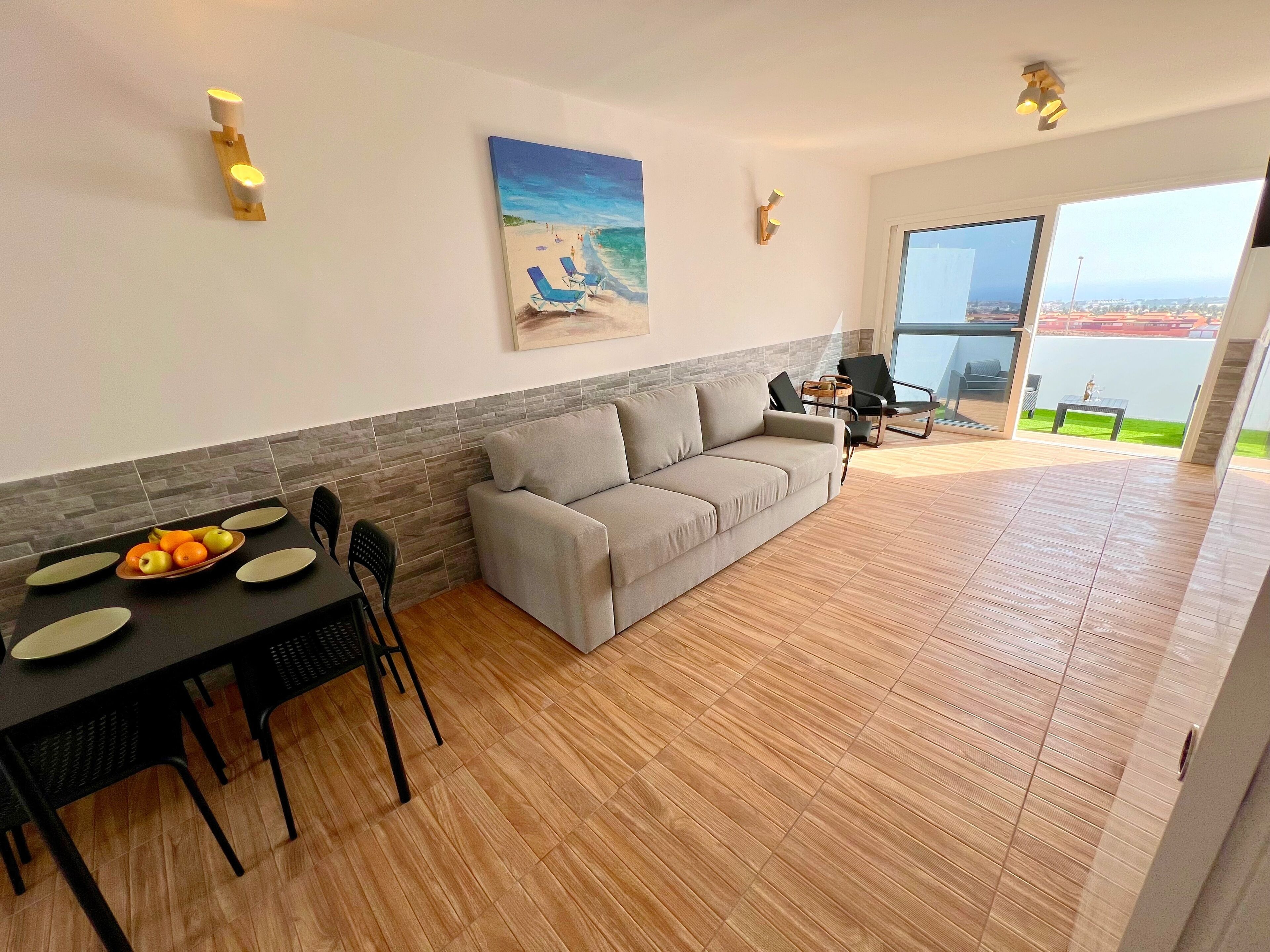 Un salon moderne avec un canapé gris, une table à manger avec de l’assiette et des fruits, et un tableau représentant une scène de plage.
