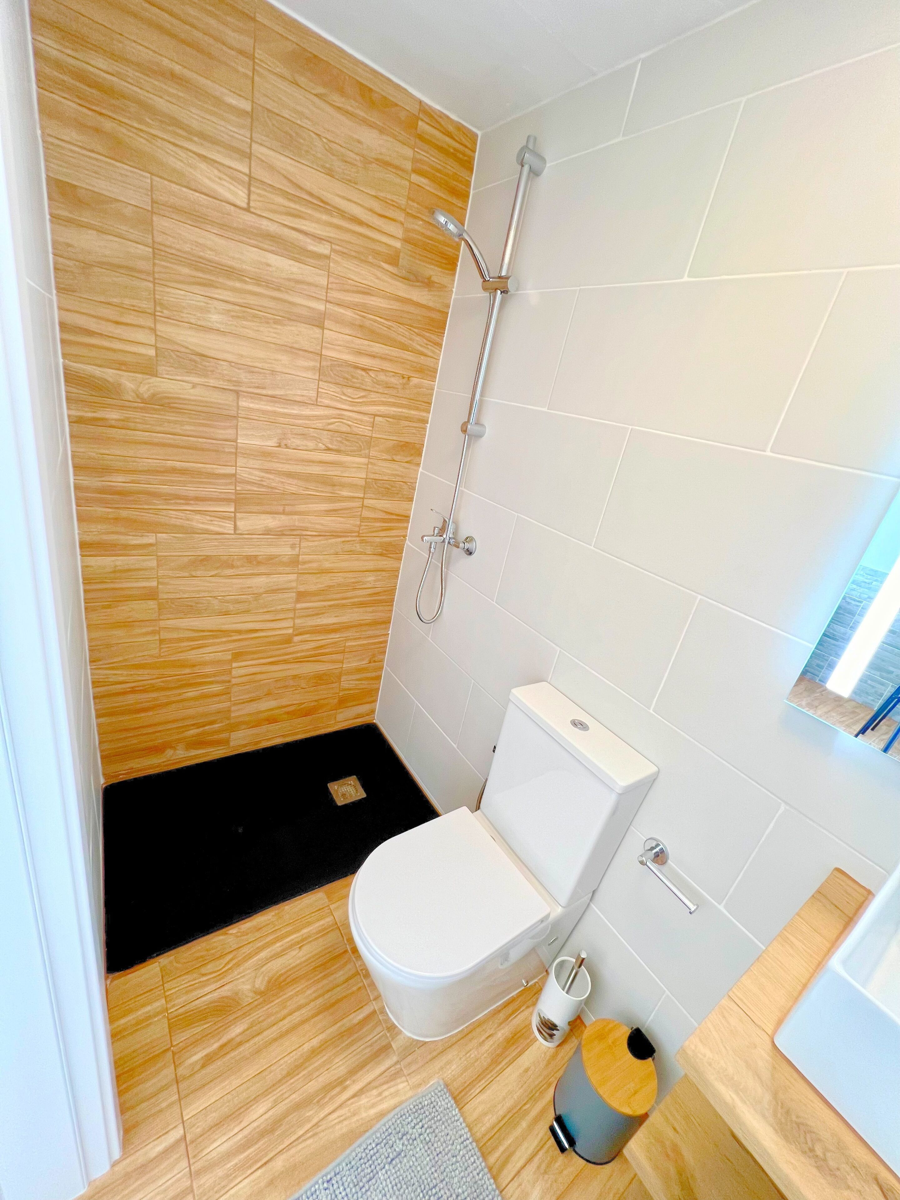 A modern bathroom with a wooden floor, white tiles, a white toilet, a shower with a handheld showerhead, and a small sink with a wooden countertop.