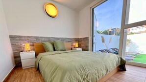 Ein Schlafzimmer mit einem großen Bett, einem Holz-Kopfteil, zwei Nachttischen mit Lampen und Blick auf eine grüne Rasenfläche durch eine Glastür.