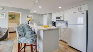 Une cuisine moderne avec des armoires blanches, un four à micro-ondes encastré et un réfrigérateur. On y trouve un petit îlot central avec un évier et un panier de fruits. La pièce est dotée d’un mur de couleur gris clair et d’un plancher en bois.