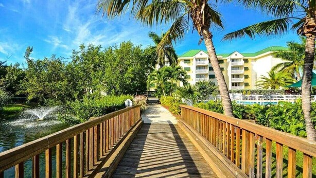 Une promenade en bois mène à un hôtel doté d’un toit végétalisé, entouré d’une végétation luxuriante et de palmiers.