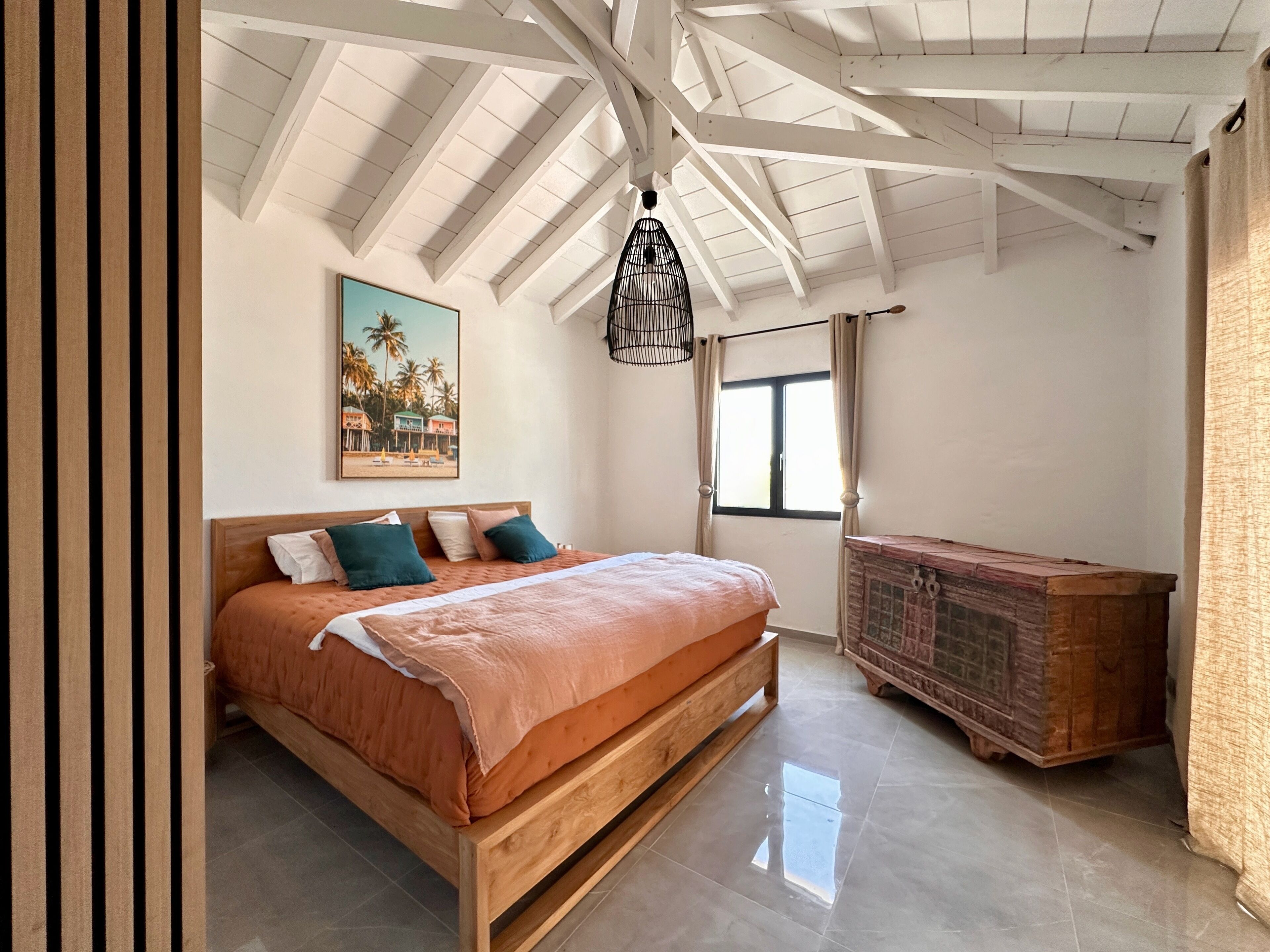 Une chambre à coucher avec un lit en bois, un tableau représentant une scène de plage, une fenêtre avec des rideaux et une armoire en bois.