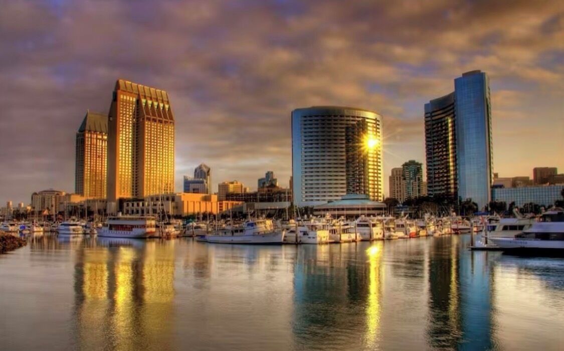 A marina with several yachts docked, a city skyline with modern buildings, and a sunset sky.