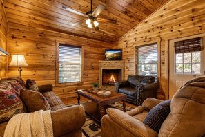 A cozy log cabin interior with a fireplace, leather furniture, and a ceiling fan.