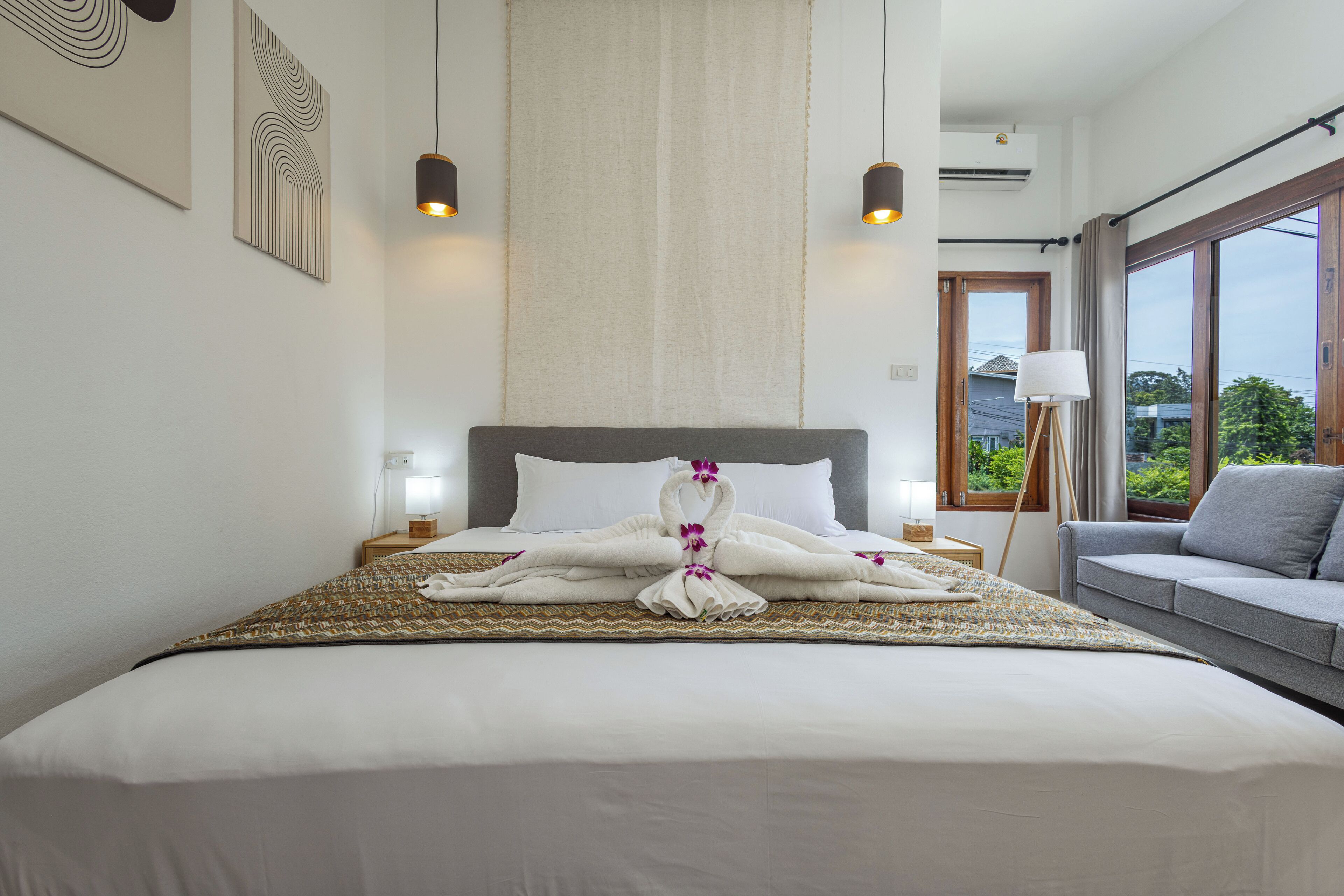 Een hotelkamer met een groot bed, een grijze bank en een raam met uitzicht op groen.