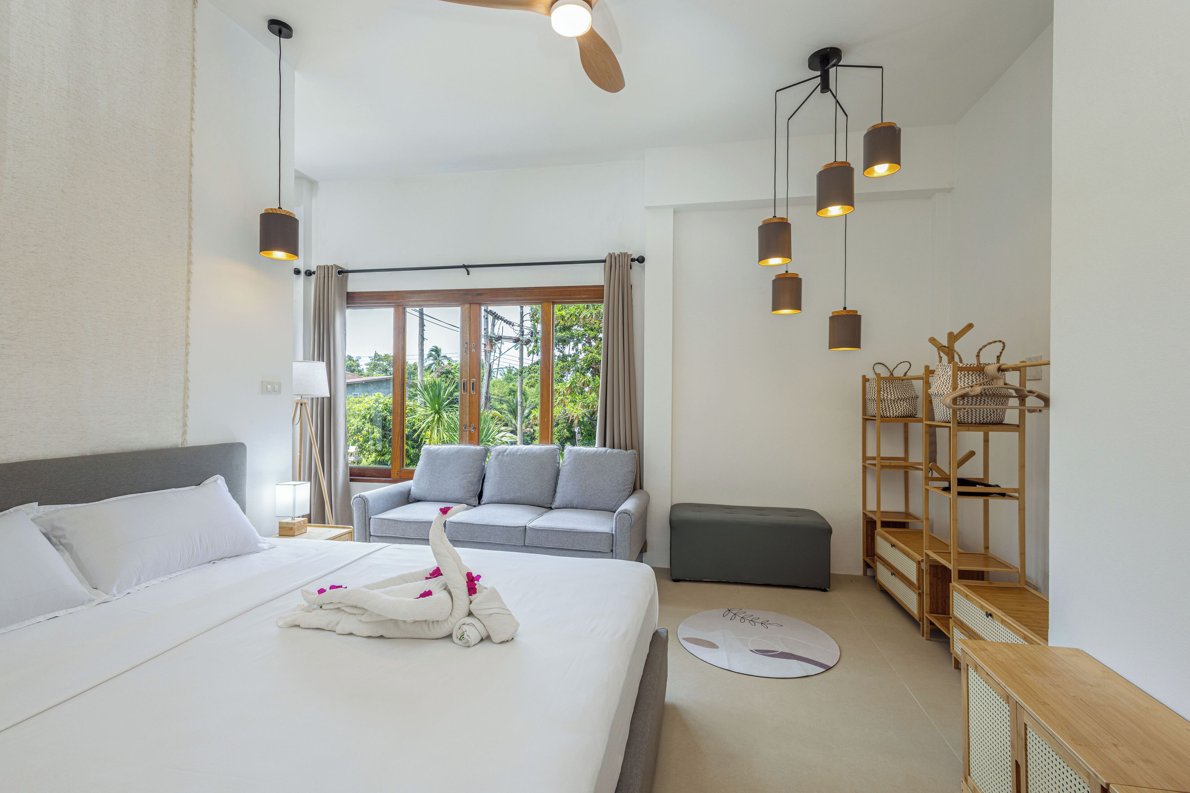 A modern hotel room with a large bed, a grey sofa, a wooden side table, and a view of greenery outside.